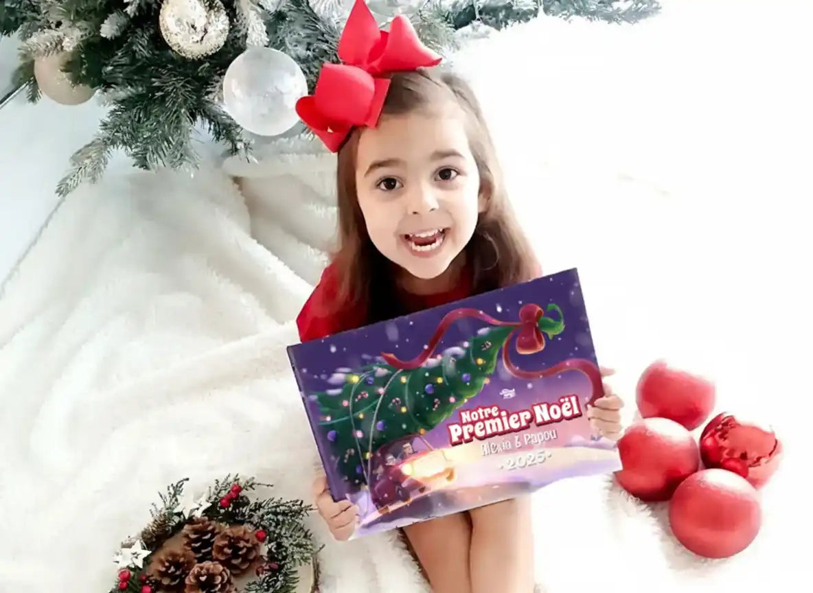 Fillette souriante tenant le livre personnalisé “Notre Premier Noël 2025” avec Alexa et Papou, assise près d’un sapin de Noël.
